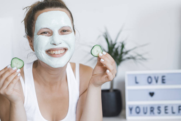 Young Pretty Blonde woman with facial mask posing with cucumber at home. Wellbeing concept, Morning skin care routine. - Foto, Imagem