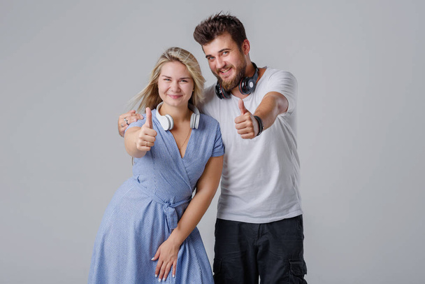 Una joven pareja alegre, un chico con una chica con auriculares inalámbricos alrededor de su cuello, en un fondo gris muestran con las manos que todo está bien
 - Foto, imagen
