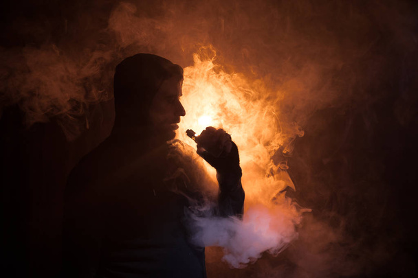 Vaping man holding a mod. A cloud of vapor. dark foggy toned background of clouds of smoke. Vaping an electronic cigarette with a lot of smoke. Vape concept. Selective focus - Photo, Image