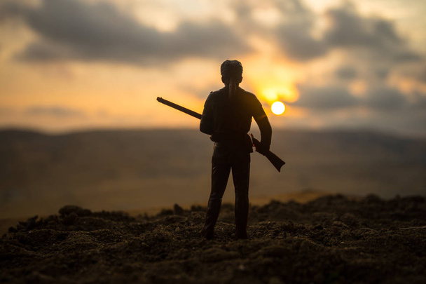 Silhouet van militaire soldaat of officier met wapens bij zonsondergang. schot, pistool, berg, kleurrijke hemel achtergrond te houden. Decoratie met speelgoed soldaat - Foto, afbeelding