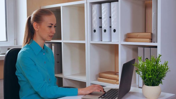 young woman has video call. worker looking on screen laptop sitting at the workspace in small office with white furniture. caucasian women with blond hair - Foto, Imagem