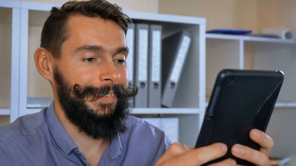 schöner Mann mit Bart und Schnurrbart mit digitalem Tablet, das lustige Fotos zeigt, die Emotionen zeigen Mitarbeiter haben Spaß mit Gadget im Büro - Foto, Bild