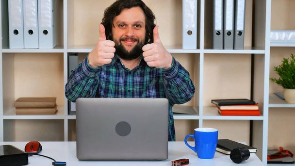 bearded man working in casual office typing on laptop showing hand gesture like. smiling worker sitting at workspace using computer - Photo, Image