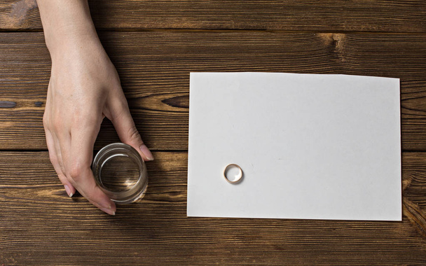 A woman's hand holds a glass with alcohol, on the table an engagement ring and a blank inscription sheet, a wooden background - Photo, image