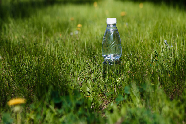 bottle of drinking water stands on the green grass outdoors in summer, blurred background - Foto, Imagen