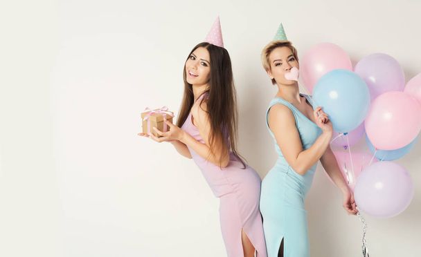 День рождения и праздник концепция. Две счастливые девушки в шапочках для вечеринок с воздушными шарами и подарками на белом фоне студии, пространство для копирования
 - Фото, изображение