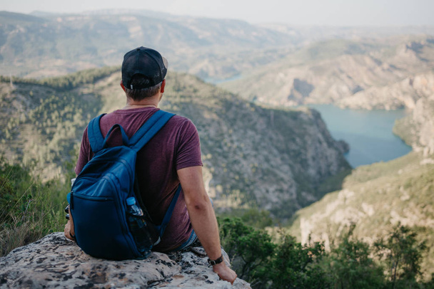 Ein Mann mit dem blauen Rucksack sitzt auf dem Stein über der wunderschönen Schlucht mit smaragdgrünem Fluss im Sonnenuntergang in Spanien. Abenteuer des Mannes. - Foto, Bild