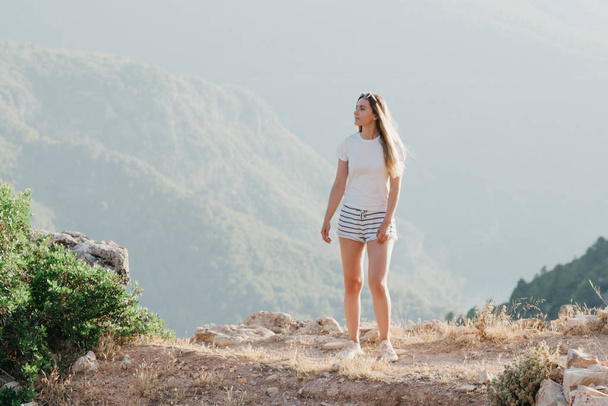 Kaunis nuori tyttö valkoisessa t-paidassa ja valkoisissa shortseissa, jotka asuvat vuorilla auringonlaskussa Espanjassa. Matkustaja vuorilla
. - Valokuva, kuva