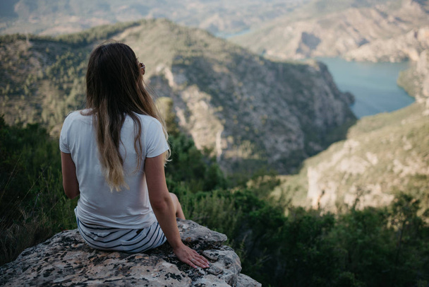 Jolie jeune fille en t-shirt blanc et short blanc avec des lunettes de soleil assis sur le rocher au-dessus du magnifique canyon avec un lac d'émeraude au coucher du soleil en Espagne. Voyageur dans les montagnes
. - Photo, image