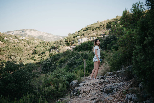 Mooie jonge meisje in het witte T-shirt en witte shorts te wachten in de bergen op de zand weg op de zonsondergang in Spanje. Reiziger in de bergen. - Foto, afbeelding