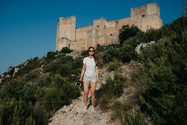Mooie jonge vrouw in het witte T-shirt en witte shorts genieten op de zand weg met het oude kasteel op de achtergrond in de bergen op de zonsondergang in Spanje. Reiziger in de bergen. - Foto, afbeelding
