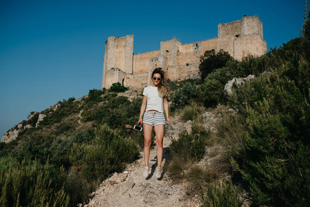 白い t シャツと白いパンツがスペインの夕暮れの砂浜道路山の背景に古代の城でジャンプではかなり若い女性。山の旅行者. - 写真・画像