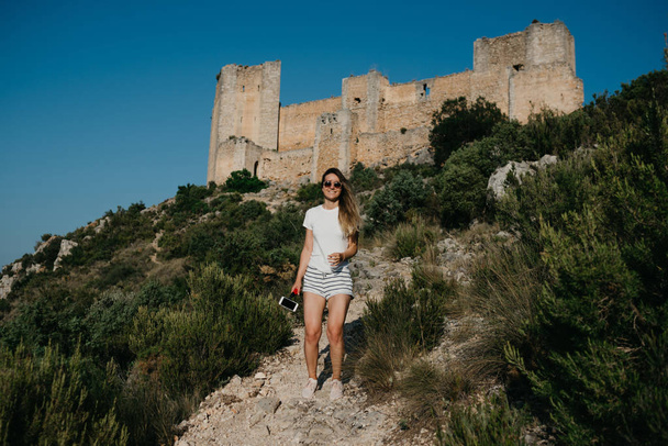 Leuke jonge vrouw in het witte T-shirt en witte shorts glimlachend op de zand weg met het oude kasteel op de achtergrond in de bergen op de zonsondergang in Spanje. Reiziger in de bergen. - Foto, afbeelding