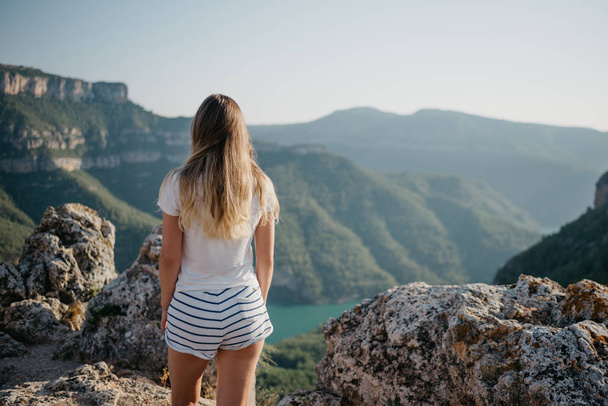 Une jolie jeune femme en t-shirt blanc et short blanc restant au sommet de la colline et regardant le canyon avec une rivière d'émeraude au coucher du soleil en Espagne. Voyageur dans les montagnes
. - Photo, image