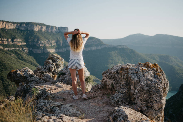 Una linda joven en camiseta blanca y pantalones cortos blancos disfrutando del cañón con un río esmeralda en la cima de la colina al atardecer en España. Viajero en las montañas
. - Foto, imagen