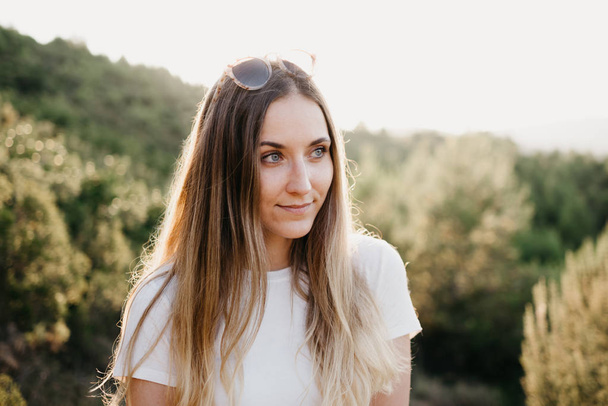 Menina bonito na camiseta e óculos de sol sorrindo entre um monte de plantas verdes na colina com o pôr do sol no fundo, na Espanha. Viajante nas montanhas
.  - Foto, Imagem
