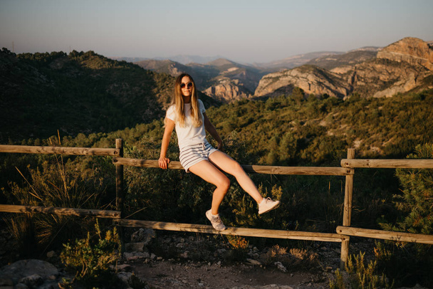 Roztomilá dívka v tričko a šortky s brýle pózuje na dřevěný plot s mnoha zelených rostlin s zelený kaňon na pozadí na západ slunce ve Španělsku. Cestovatel v horách. - Fotografie, Obrázek