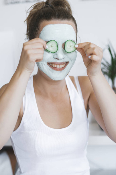 Young Pretty Blonde woman with facial mask posing with cucumber at home. Wellbeing concept, Morning skin care routine. - Photo, Image