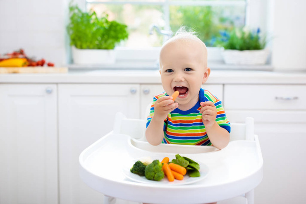 Bébé mignon manger des légumes dans la cuisine blanche. Le sevrage des nourrissons. Petit garçon essayant des aliments solides, brocoli biologique, chou-fleur, carotte et pois verts. Une alimentation saine pour les enfants. Enfant mordant la carotte
.  - Photo, image