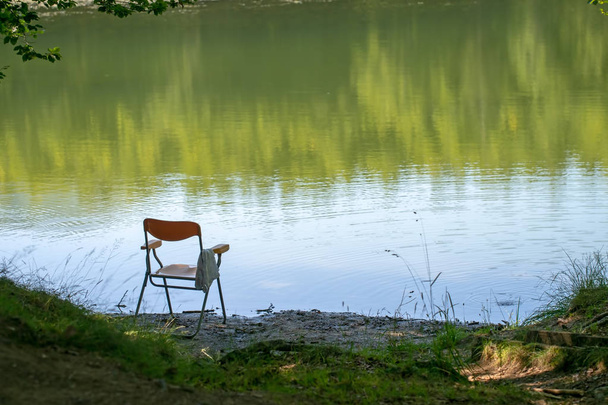 Angeln gegangen, vielleicht, Stuhl am Seeufer zurückgelassen. Sommer. Im Hintergrund sitzt niemand auf der Bank. - Foto, Bild
