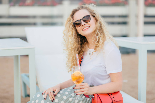 Aantrekkelijke jonge vrouw met golvend haar, zonnebril draagt, heeft goede rust tijdens zomertijd, houdt van frisse drank, heeft vriendelijke glimlach, kijkt positief naar de camera. Mensen, drinken, recreatie-concept - Foto, afbeelding