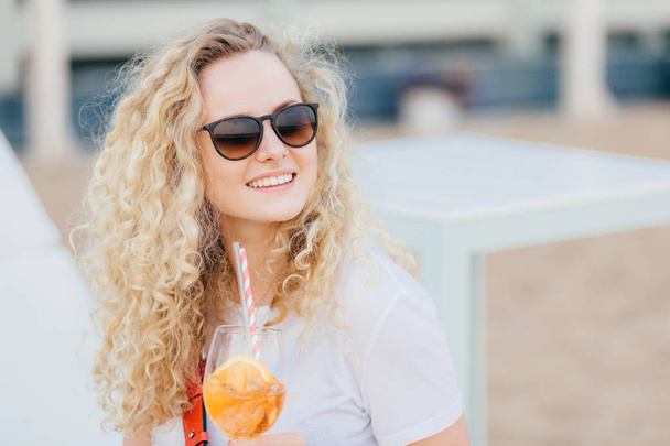 Cheerful blonde female with wavy light hair, positive smile, wears shades, looks aside with happy expression, drinks fresh cocktail, poses against blurred background with copy space for advertisement - Photo, Image