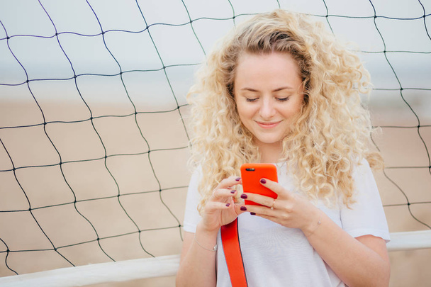 Mladá žena má kudrnaté vlasy drží moderní chytrý telefon stahuje fotky ze sociálních sítí, představuje čisté pozadí tenis, využívá internet zdarma. Krásná blondýnka využívá elektronické zařízení - Fotografie, Obrázek