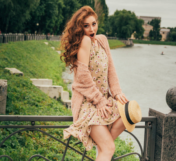 beautiful red-haired girl.picture against the background of the river, water, lake in the park, summer..sits on a fence, in the hands of a hat, a beige dress in a flower and a beige sweater.the wind raises the skirt of the dress, the girl is surprise - Photo, image