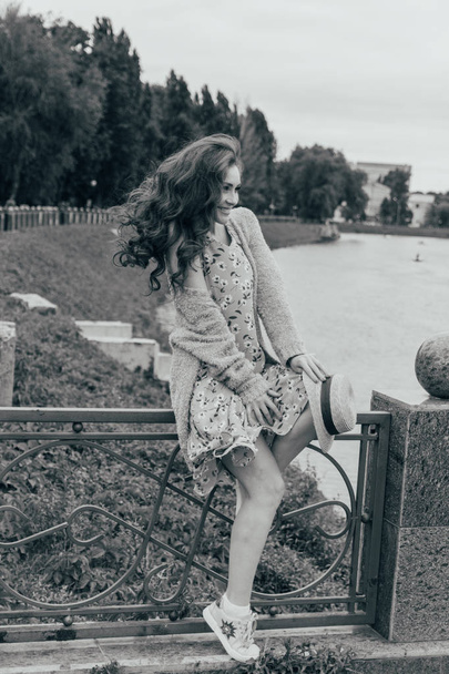 sits on a fence, in the hands of a hat, a dress in a flower and a sweater.beautiful girl.picture against the background of the river, water, lake in the park, summer.hair and dress evolve from the wind.black and white photo - Zdjęcie, obraz