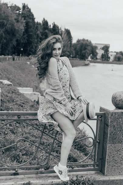 sits on a fence, in the hands of a hat, a dress in a flower and a sweater.beautiful girl.picture against the background of the river, water, lake in the park, summer.hair and dress evolve from the wind.black and white photo - Fotografie, Obrázek