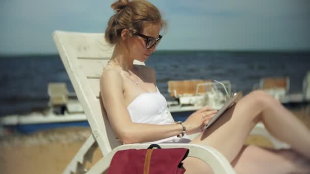 A young girl in a white bikini lies and tans on a deckchair on a sea sandy beach and is working on a tablet - Felvétel, videó