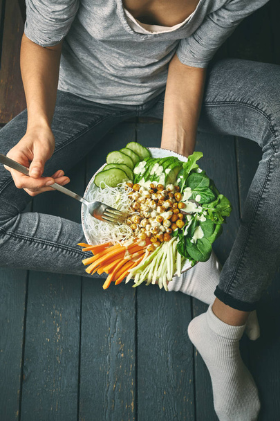 top view of Healthy breakfast, raw vegan food in Buddha Bowl, fried chickpeas, rice noodles with raw wegetables and spinach. Woman in grey jeans holding fork and bowl with legs and hands visible, view - Fotó, kép