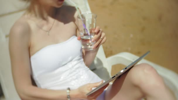 Beyaz bikinili genç bir kız yatıyor ve tans üzerinde bir deniz kumlu plajda şezlong ve bir tablet üzerinde çalışan - Video, Çekim