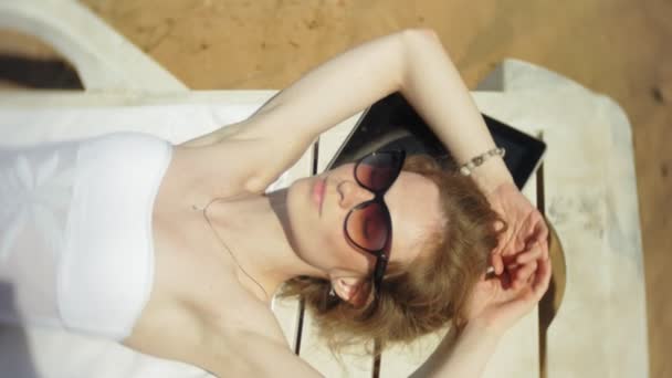 Una giovane ragazza in bikini bianco si sdraia e abbronza su una sdraio su una spiaggia di sabbia marina e sta lavorando su un tablet
 - Filmati, video
