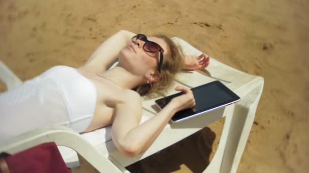 Beyaz bikinili genç bir kız yatıyor ve tans üzerinde bir deniz kumlu plajda şezlong ve bir tablet üzerinde çalışan - Video, Çekim