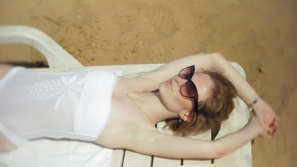 Nuori tyttö valkoisissa bikineissä makaa ja ottaa aurinkoa kansituolilla meren hiekkarannalla
 - Materiaali, video