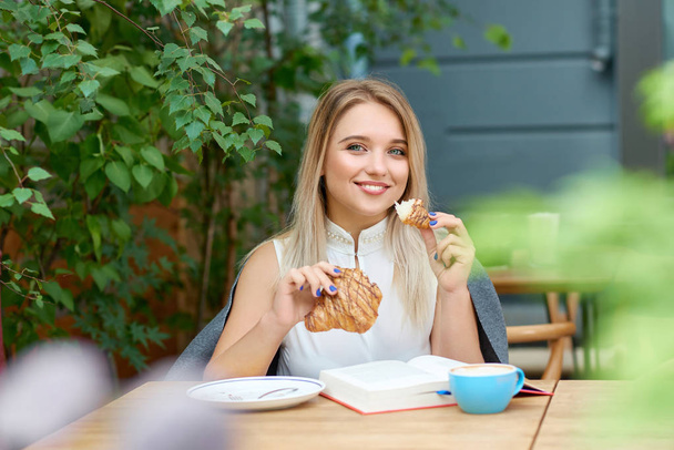 Smiling girl with blonde hair eating croissant, looking at camera. - Fotó, kép
