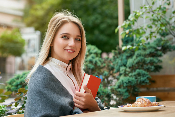 Cute girl holding big red book sitting outdoors cafe, eating croissant. - Фото, изображение
