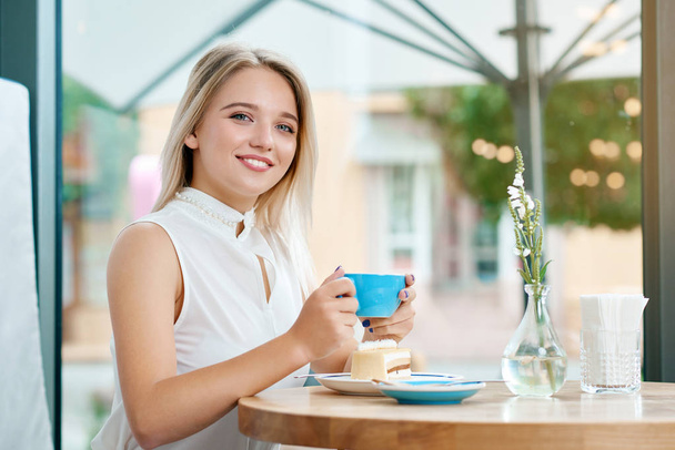 Cute blonde girl keeping cup of coffee on outdoors, smiling. - Foto, Imagem
