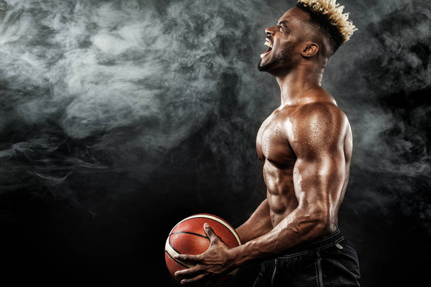 Portrait of afro-american sportsman, basketball player with a ball over black background. Fit young man in sportswear holding ball. - Foto, imagen