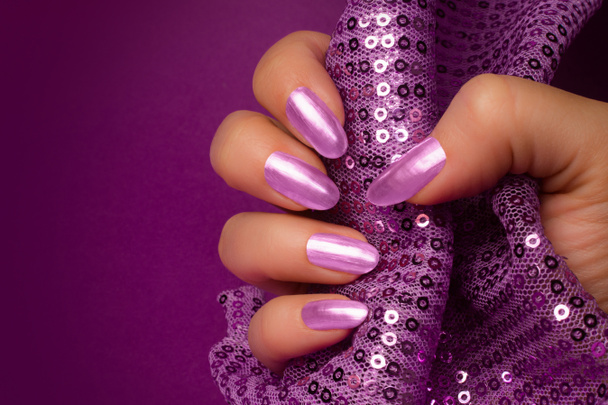 Female hand with shiny purple nails is holding purple glittered fabric on purple background, nail care and manicure concept. - Photo, Image