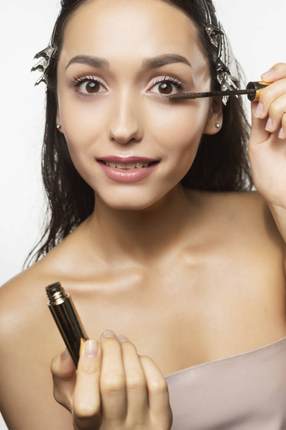 Beautiful young brunette girl model with naked shoulders and wet hair with hair grips, nude makeup puts mascara on her lashes on white background. Healthy clean skin. Advertising and commercial design - 写真・画像