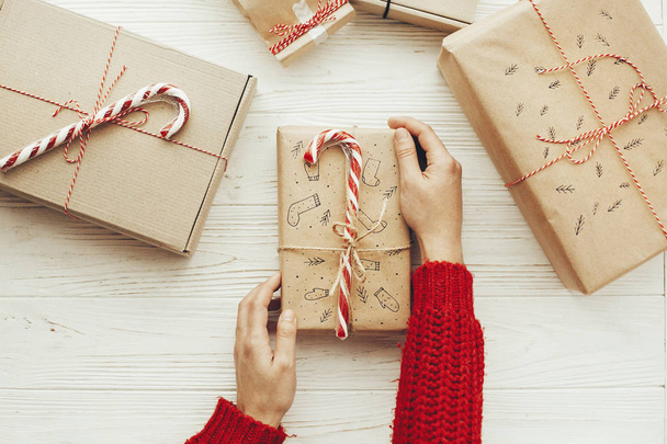 Frauenhände halten ein stilvolles Weihnachtsgeschenk mit Zuckerrohr und roter Schleife in der Hand. flach lag. moderne handwerkliche Geschenke auf weißem Holzgrund. Saisongrüße. Frohe Weihnachten - Foto, Bild