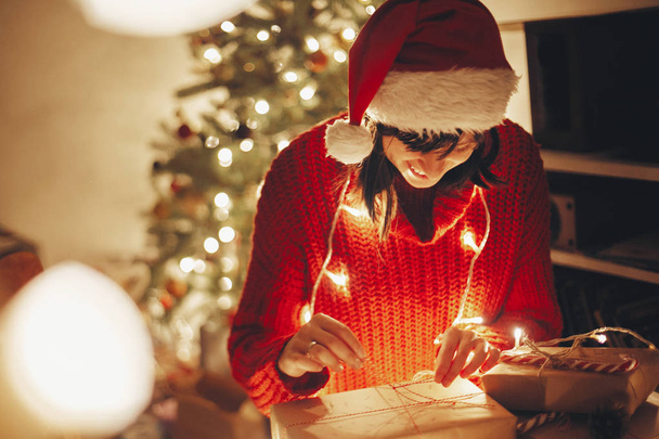 girl in santa hat and red sweater wrapping christmas presents in lights in evening festive room under tree illumination. kid opening modern craft gifts with candy cane. atmospheric moments - Photo, Image