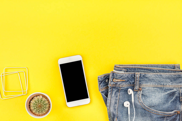 Plochá laických modré džíny, kaktus a smartphone sluchátka na odvážné žluté pozadí s kopie prostoru. Pohled z ženy ležérní oblečení. Módní bokové vypadat pohled shora - Fotografie, Obrázek