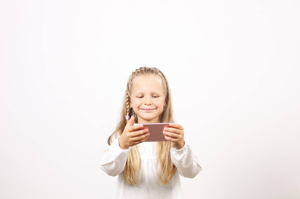 Portrait of positive & funny four year old blonde little girl, holding mobile cell phone, smiling at camera, taking selfies. Childhood & technology concept. Isolated background, copy space, close up. - Foto, Imagem