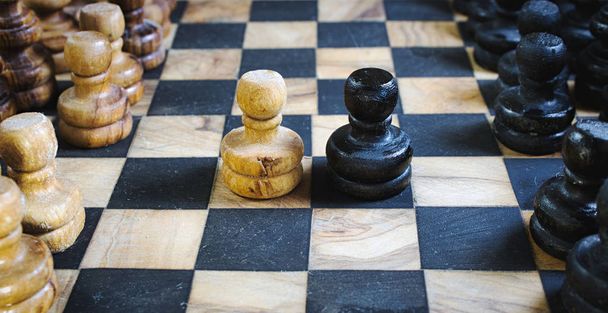 Antiguo tablero de ajedrez de madera de olivo con piezas acérrimas y peones blancos y negros luchando cabeza a cabeza
 - Foto, imagen