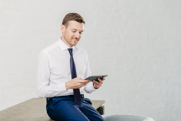 Positive male financier wears white shirt, tie, sits on table, installs new application on touch pad, connected to wireless internet. Successful businessman reads notification on modern device - Фото, зображення