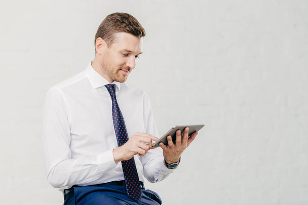 Positive successful male executive manager holds modern touch pad, gets notification about net feeds in social networks, satisfied with high speed internet connection, dressed in formal shirt with tie - Foto, Bild