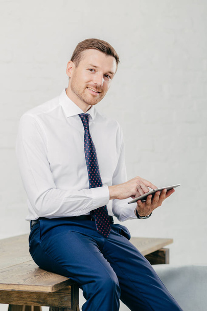 Smiling young businessman in elegant clothes happy to recieve income message on modern tablet computer, sits at wooden office desk in his cabinet, poses against white wall. Business concept. - Photo, Image